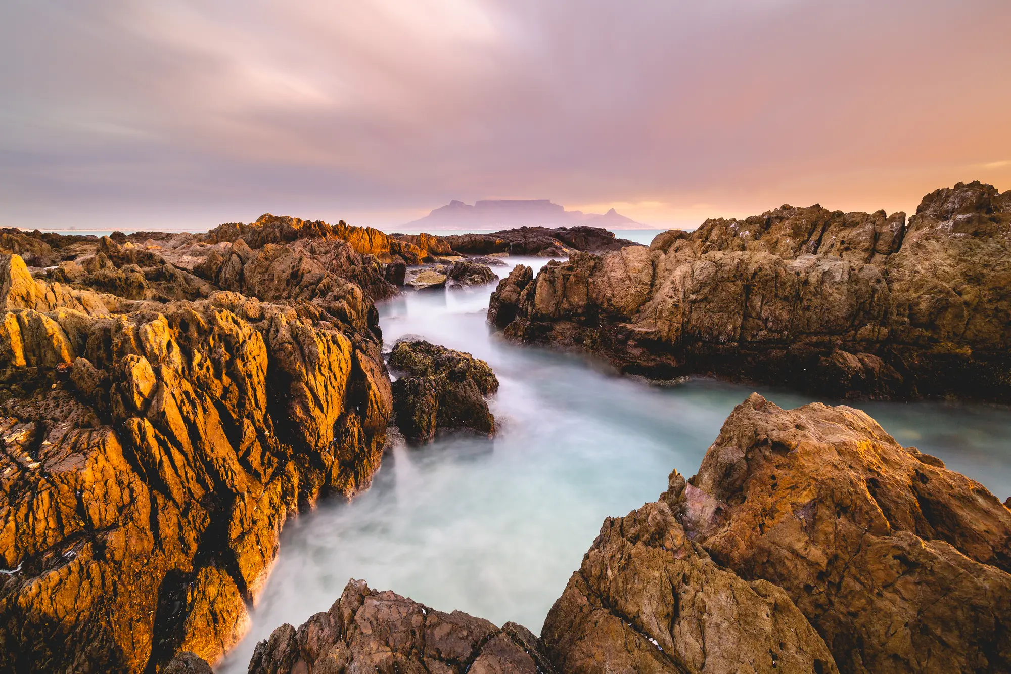 Long-exposure seascape from the rocks at Blouberg, silky water pooling between jagged rock formations with Table Mountain under a dusty pink sky