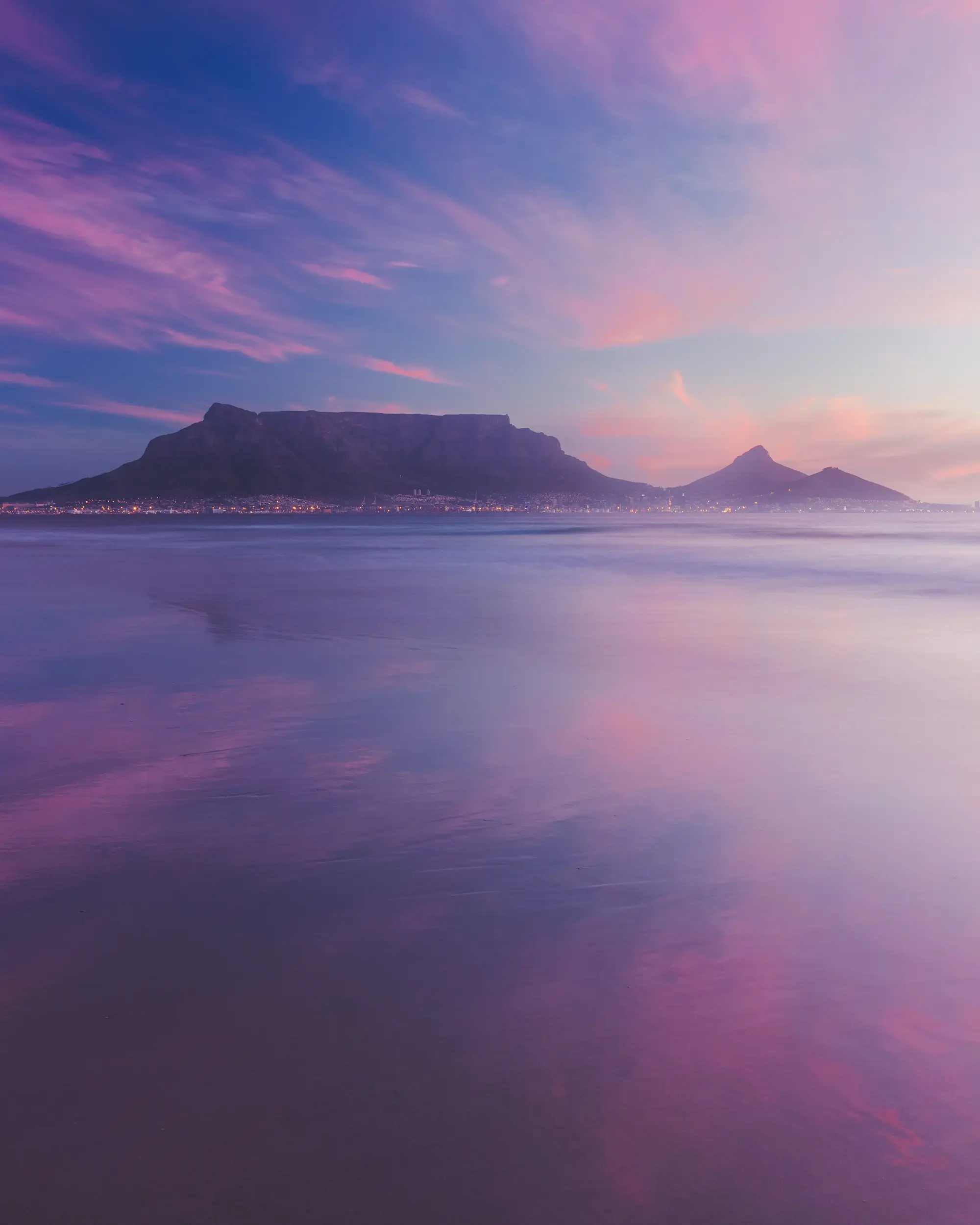 Table Mountain and Lion's Head reflected in glassy shallows at Blouberg, the sunset sky dissolving into violet and pink
