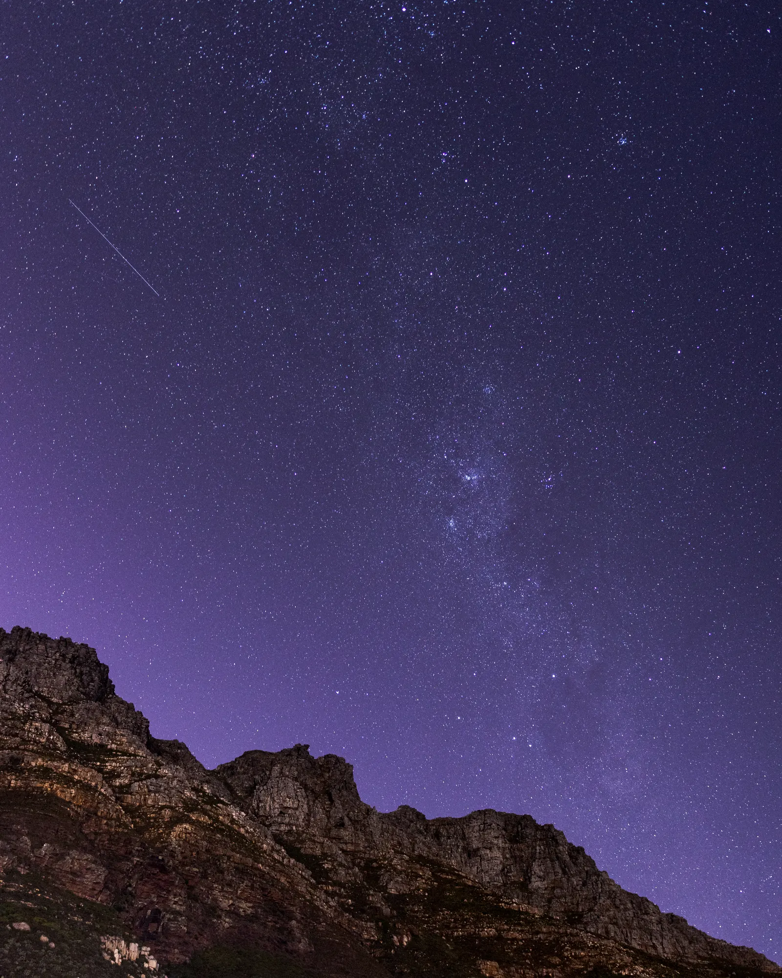 The Milky Way rising above the jagged ridgeline of the Twelve Apostles, a satellite trail crossing the deep violet sky