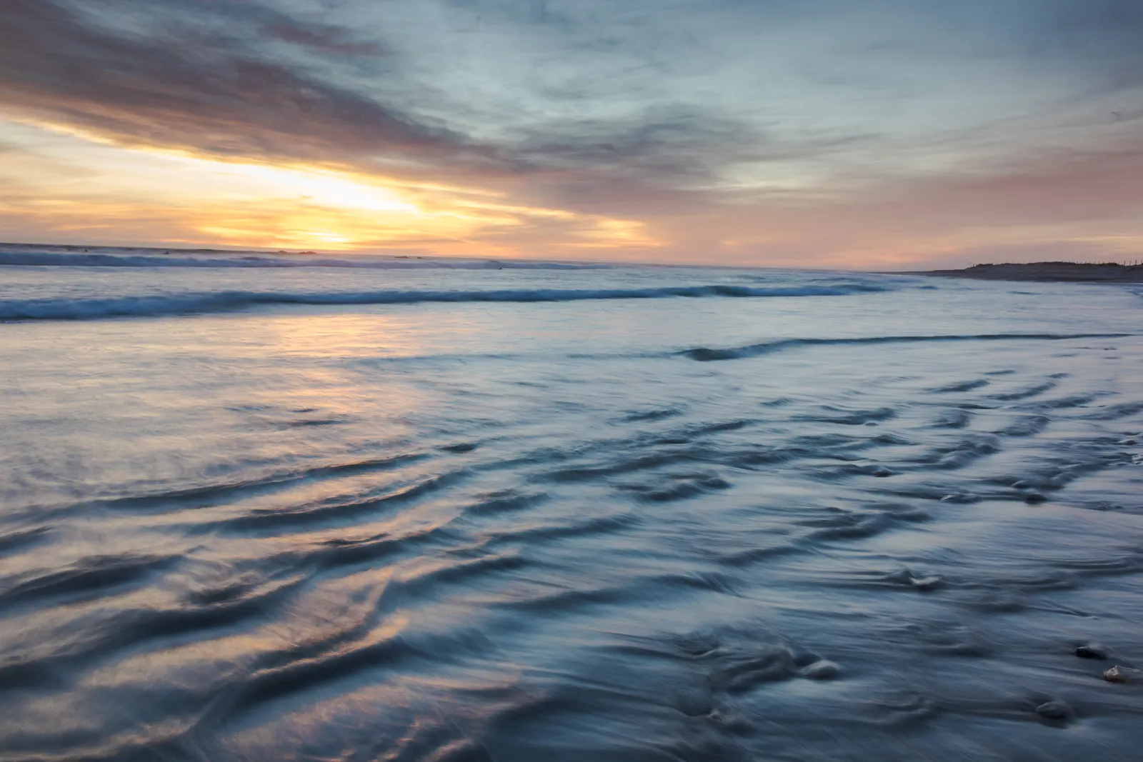 Water rushing back across sand and rock at sunset, a gold horizon under a soft grey sky