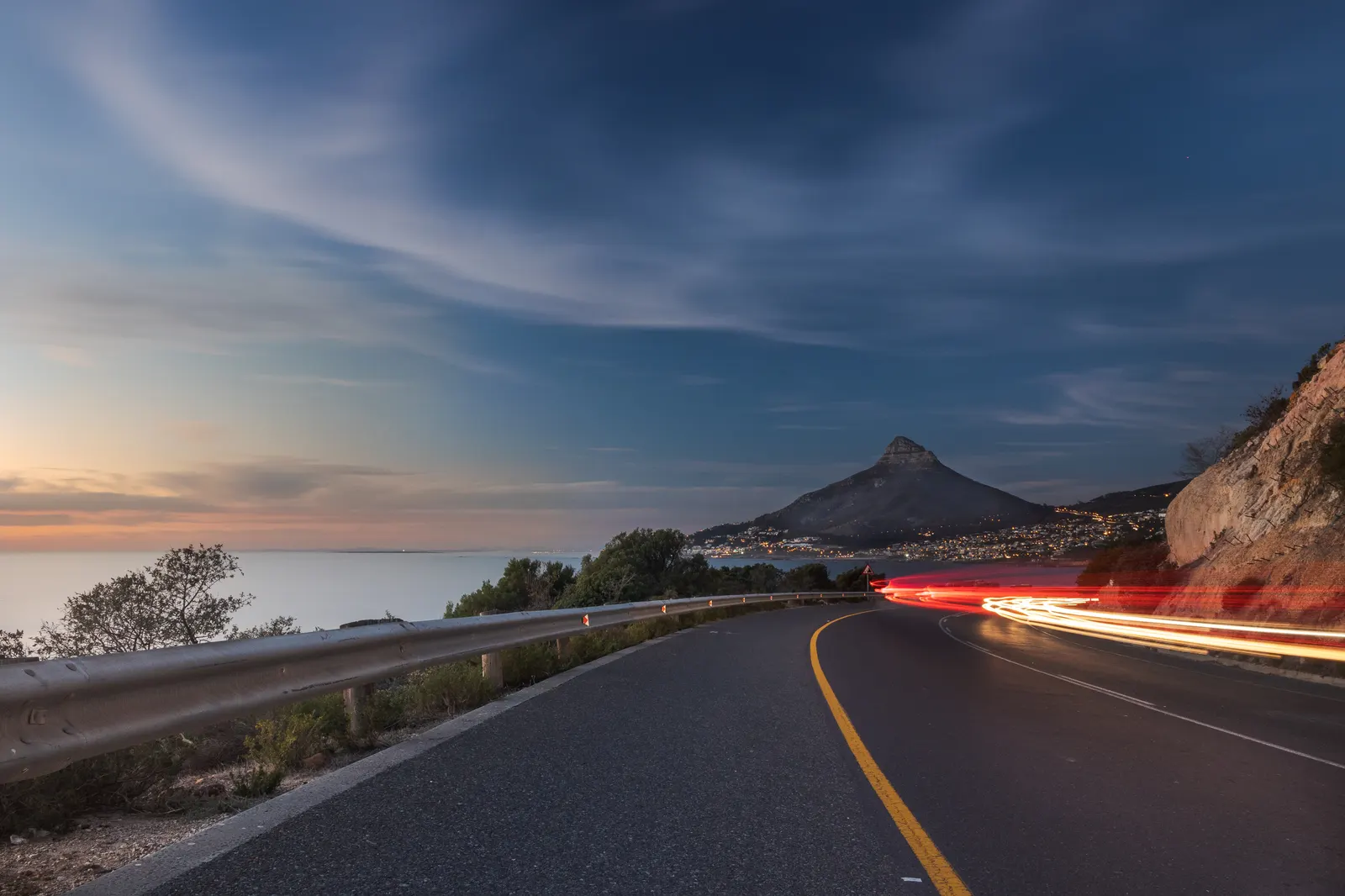 Car light trails sweeping around the bend on Victoria Road at blue hour, Lion's Head rising beyond the bay and Camps Bay glowing below