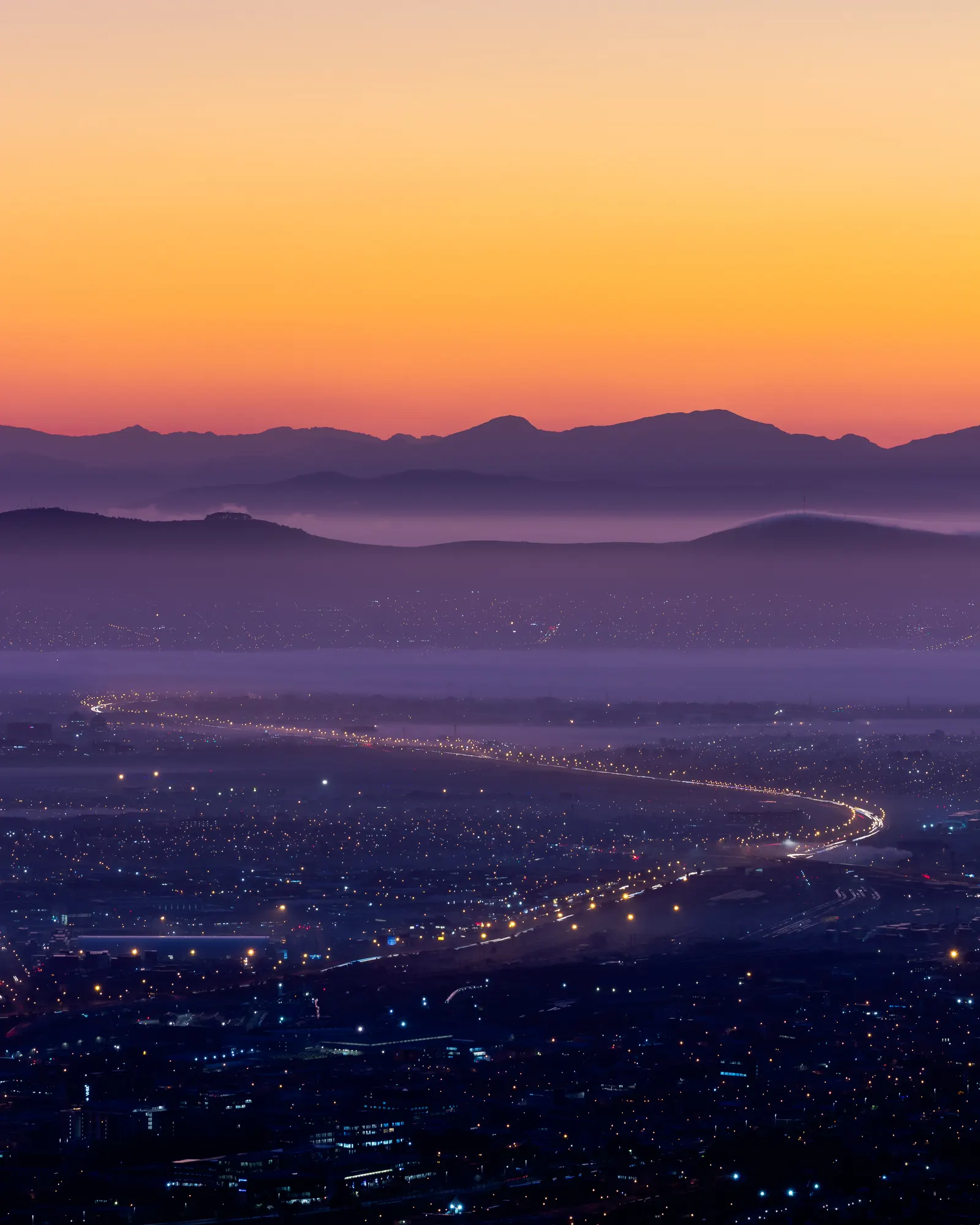 The N1 highway tracing a luminous S-curve toward Cape Town in the pre-dawn dark, layers of mountain and mist toward an orange horizon
