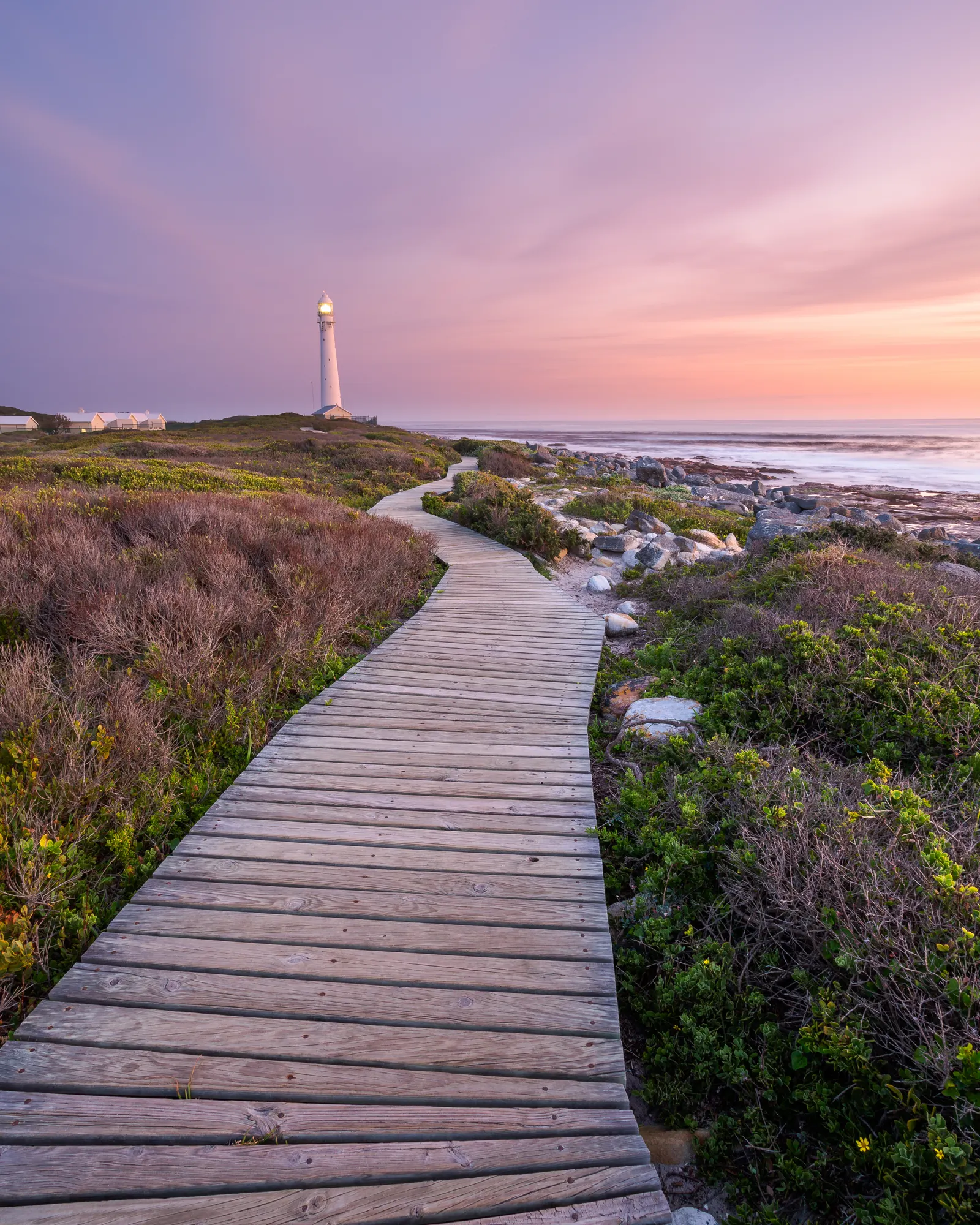 Winding boardwalk through fynbos leading toward Slangkop Lighthouse at dusk under a warm pastel sky