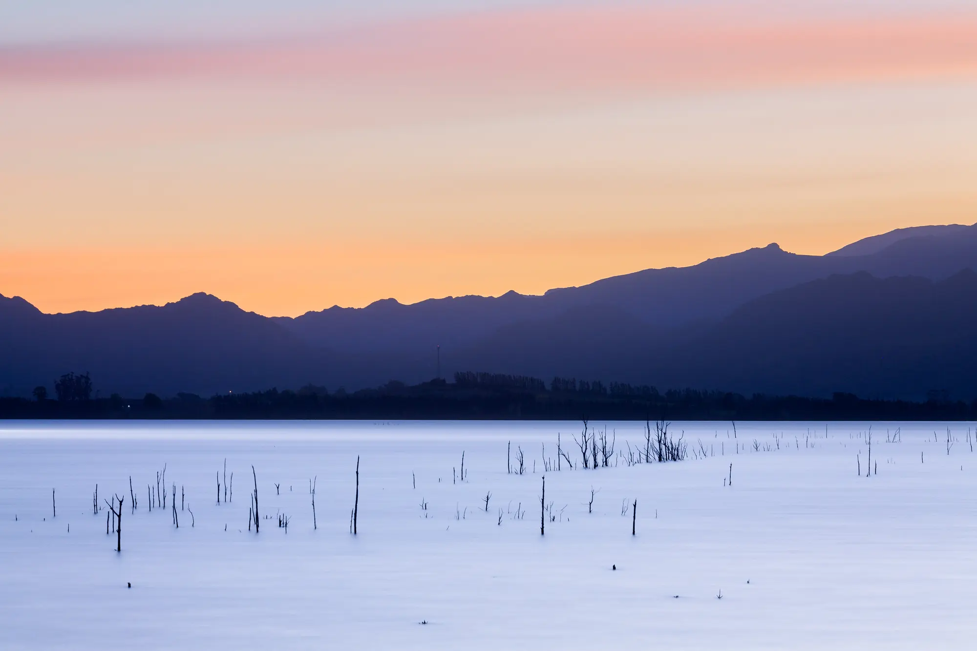 Dead tree skeletons rising from the still surface of Theewaterskloof Dam at sunset, mountains layered behind a burning orange horizon