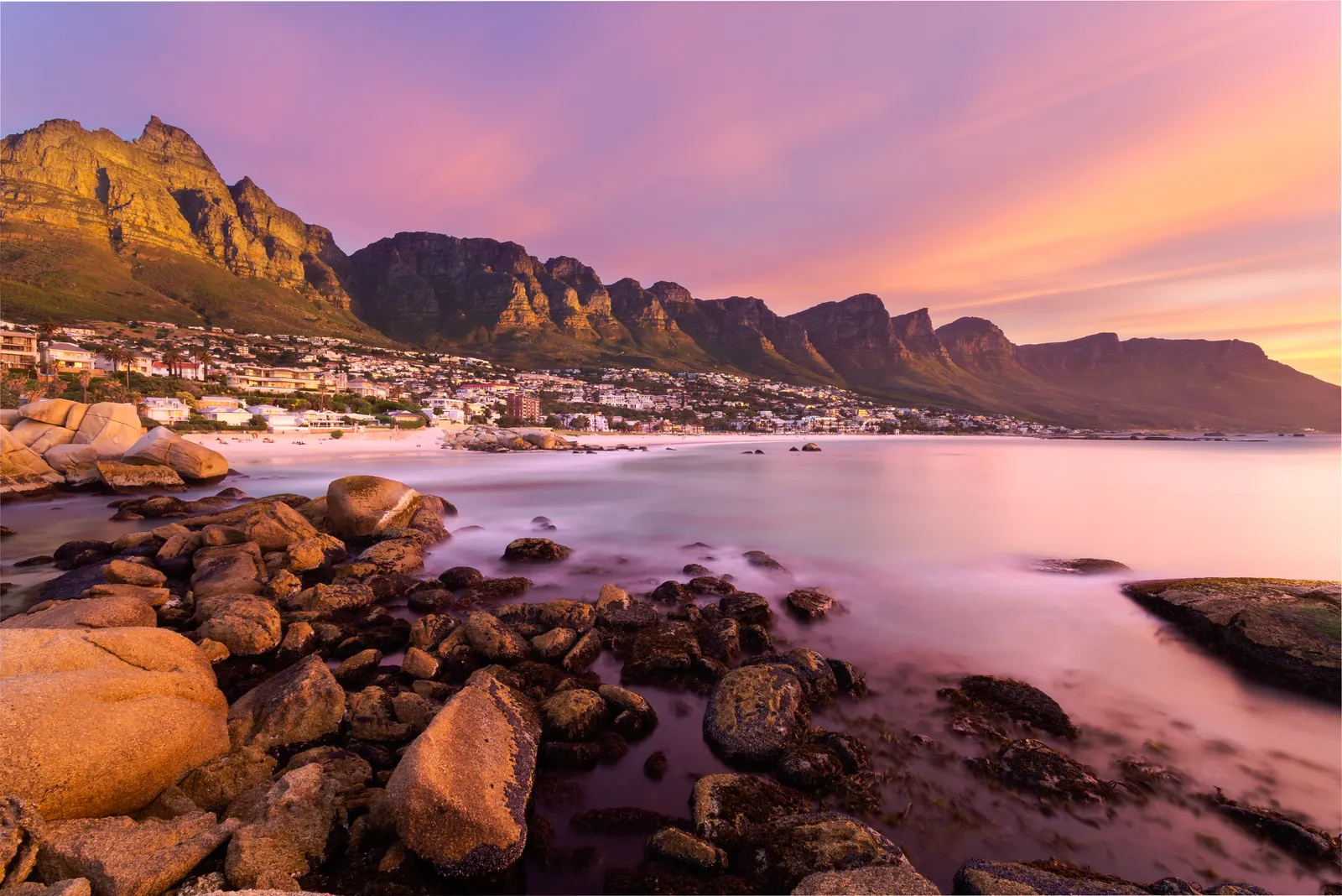 The Twelve Apostles mountains lit gold at sunset from Maiden's Cove, granite boulders and silky water in the foreground under a pink and orange sky
