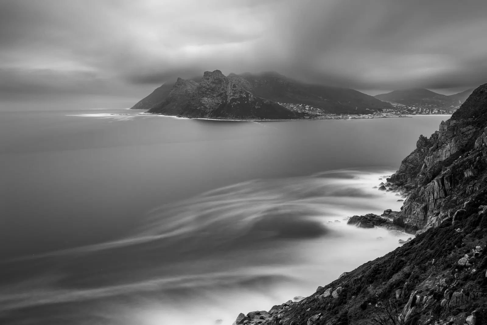 Monochrome long exposure of Hout Bay from Chapman's Peak, cloud-wrapped mountains above a smoothed, swirling sea