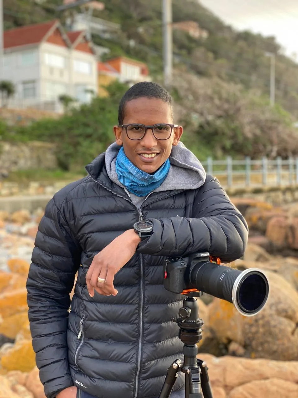Keagan De Villiers on location at the coast, smiling beside his camera and tripod among the rocks